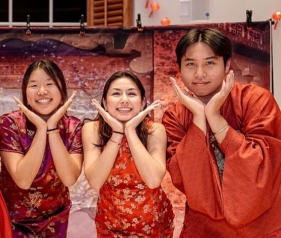 Three young adults dressed in traditional Asian attire smile and pose with their hands under their chins in a playful gesture, standing in front of a festive backdrop with warm lighting.