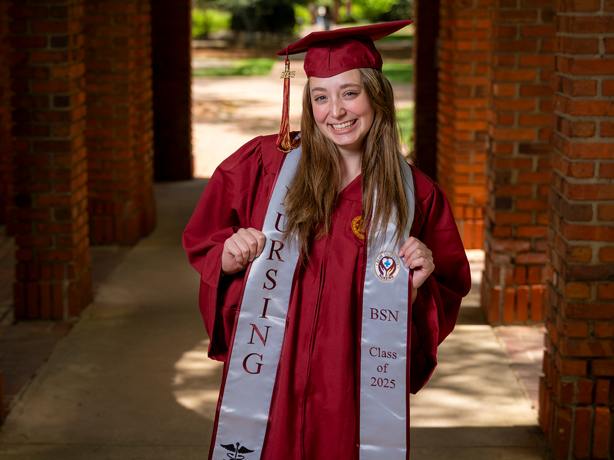 A smiling nursing graduate in a maroon cap and gown holds the ends of her stole that reads “NURSING BSN Class of 2025” while standing in a brick archway.