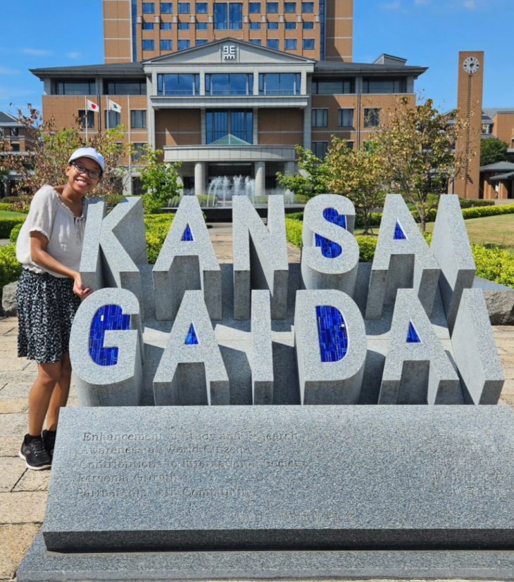 A smiling person in a white cap poses beside a large stone sign that reads “KANSAI GADAI” in bold letters, with a university building and fountain in the background on a sunny day.