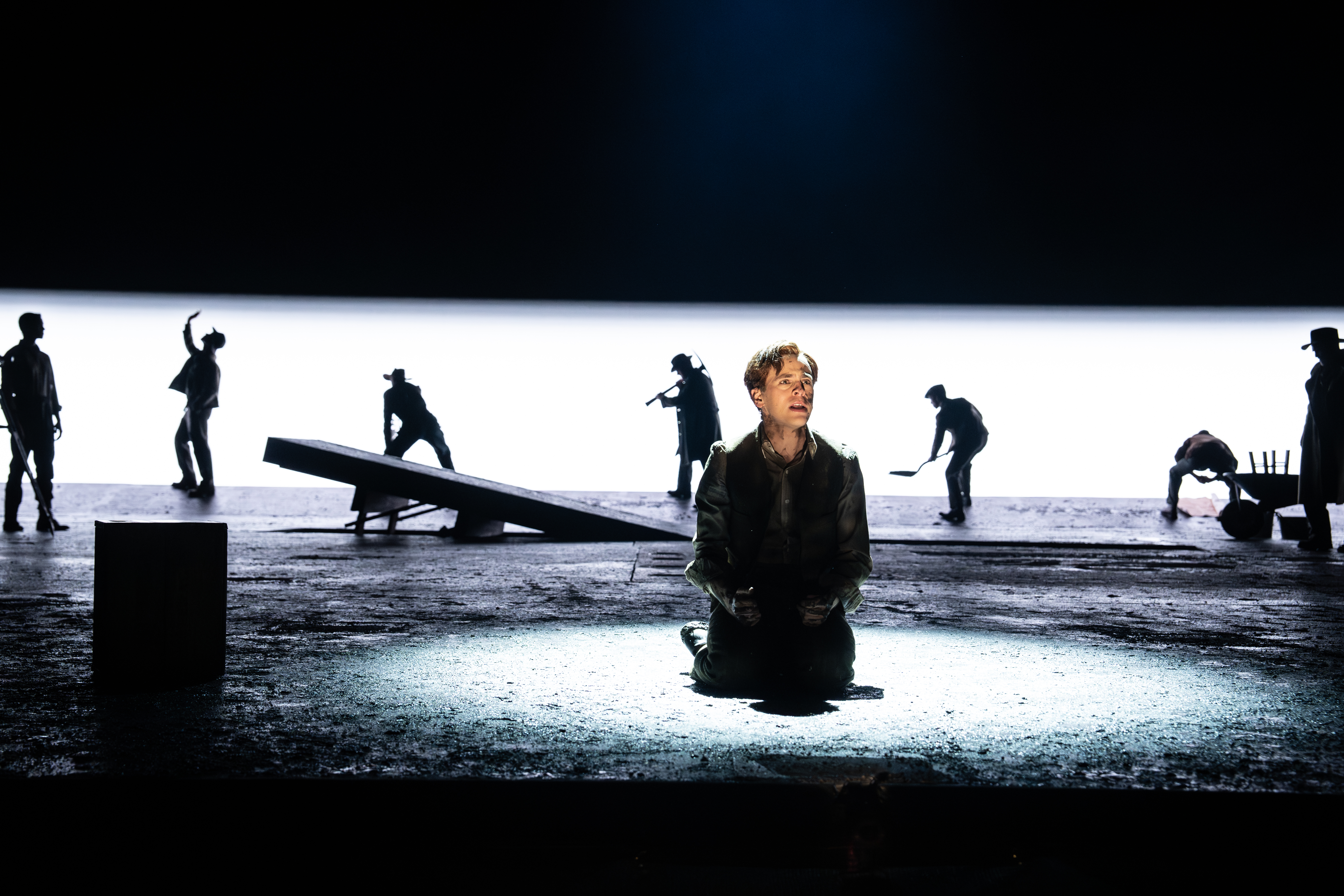 An actor kneels in a spotlight at center stage while silhouetted performers move dynamically behind him against a stark white background.