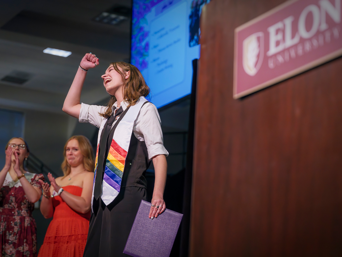 A student wearing a rainbow stole raises their fist and smiles proudly on stage at an Elon University event, as others applaud in the background.