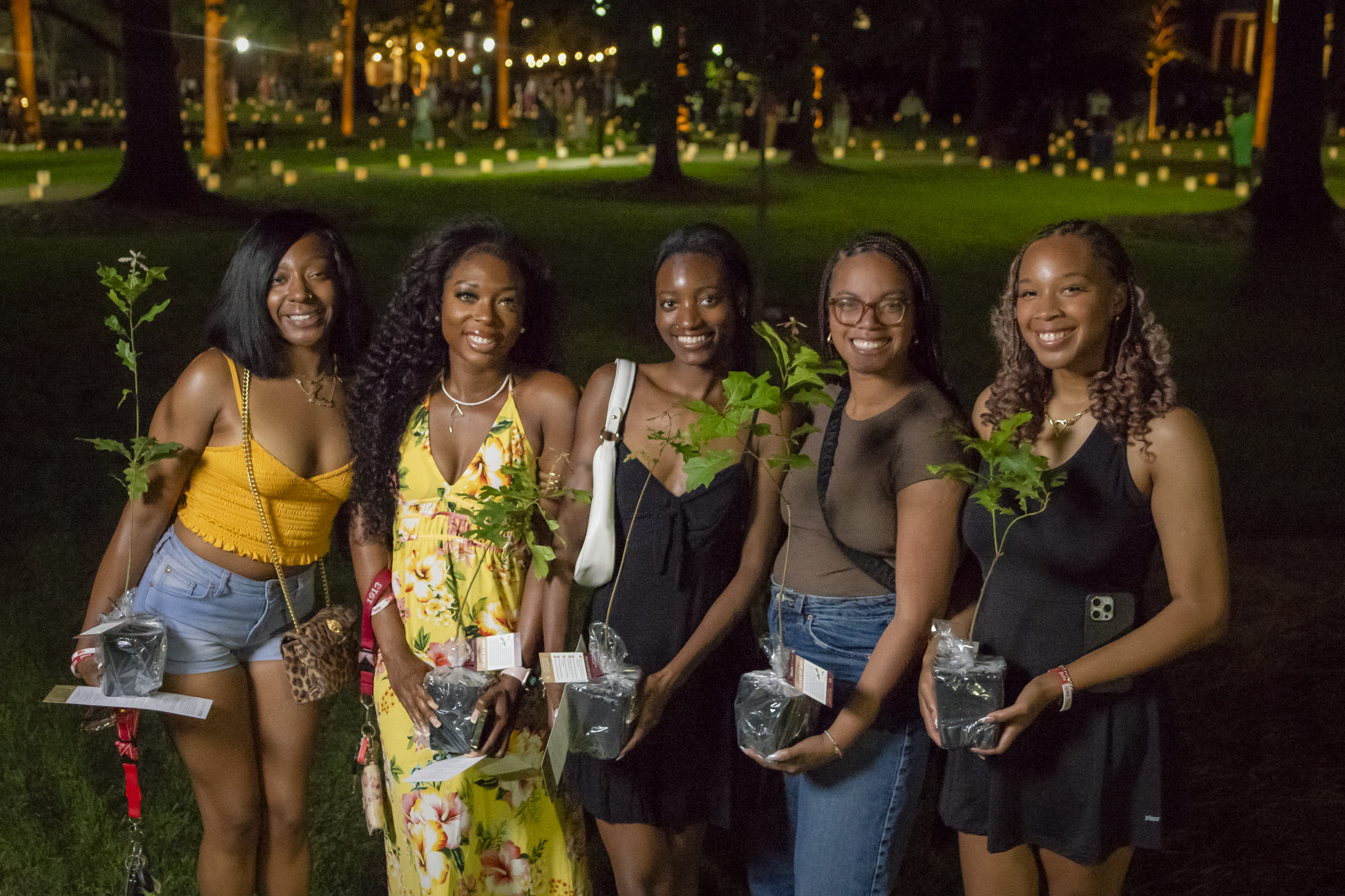 Five graduating seniors smile while holding oak saplings during Elon’s Numen Lumen ceremony, symbolizing their growth since receiving acorns as first-year students.