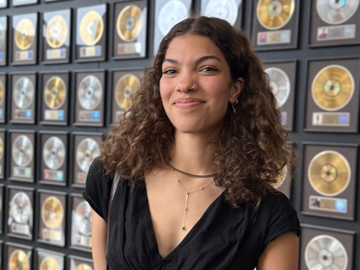 A young woman with curly hair smiles confidently in front of a wall filled with framed gold and silver records.