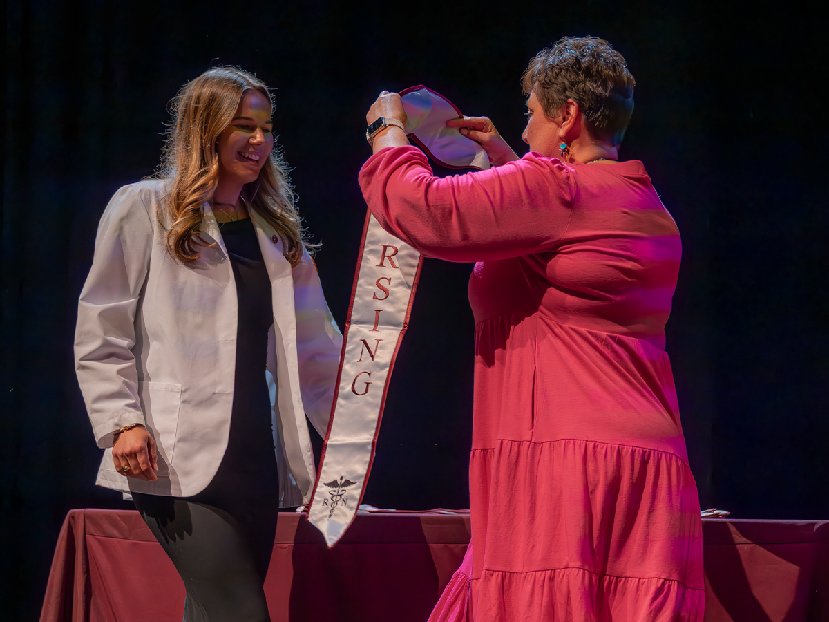 The inaugural cohort of Elon’s Bachelor of Science in Nursing program poses in white coats and stoles on the steps outside a campus building.