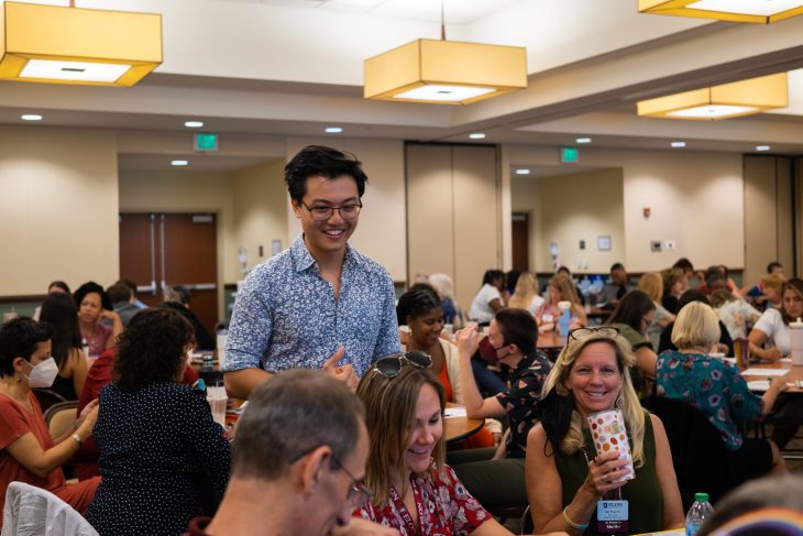A smiling facilitator stands among attendees engaged in lively conversation during a professional workshop held in a large, well-lit conference room.