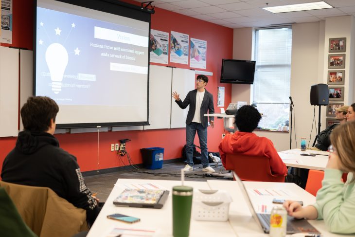 A student presents in front of a projected slide in a brightly lit classroom as peers seated around tables listen and take notes.