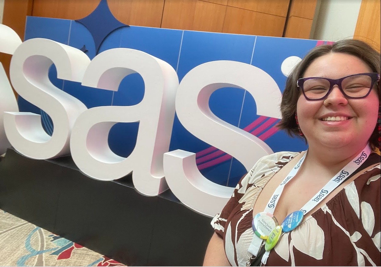 A smiling attendee takes a selfie in front of a large “SAS” sign at a conference, wearing a name badge and lanyard.