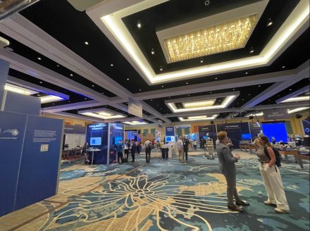 Attendees walk and network among booths and displays inside a brightly lit, modern conference hall with a patterned carpet and chandeliers.