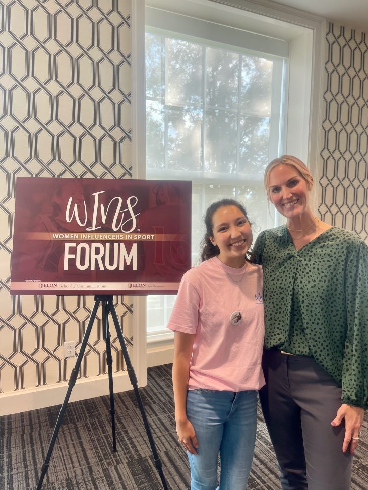 Two women smile and pose together next to a sign for the “Women Influencers in Sport Forum” hosted by Elon University’s School of Communications.