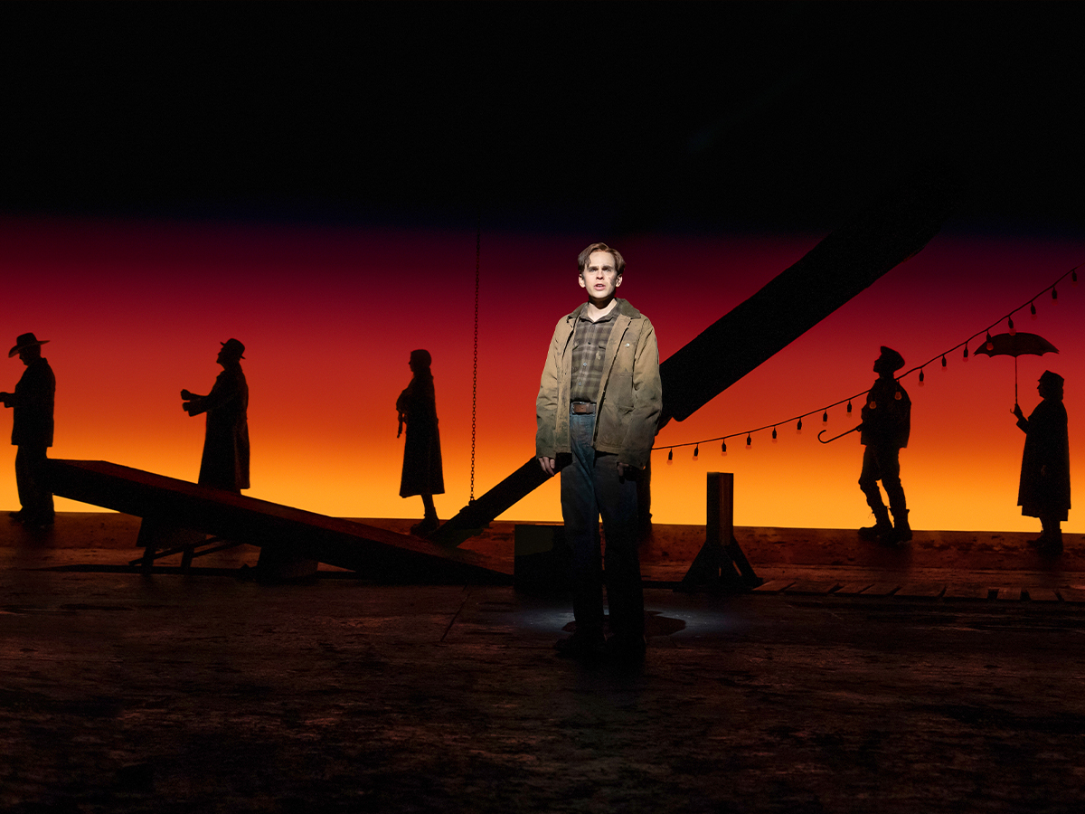An actor stands in a spotlight on stage as silhouetted figures move behind him against a vivid sunset-colored backdrop.