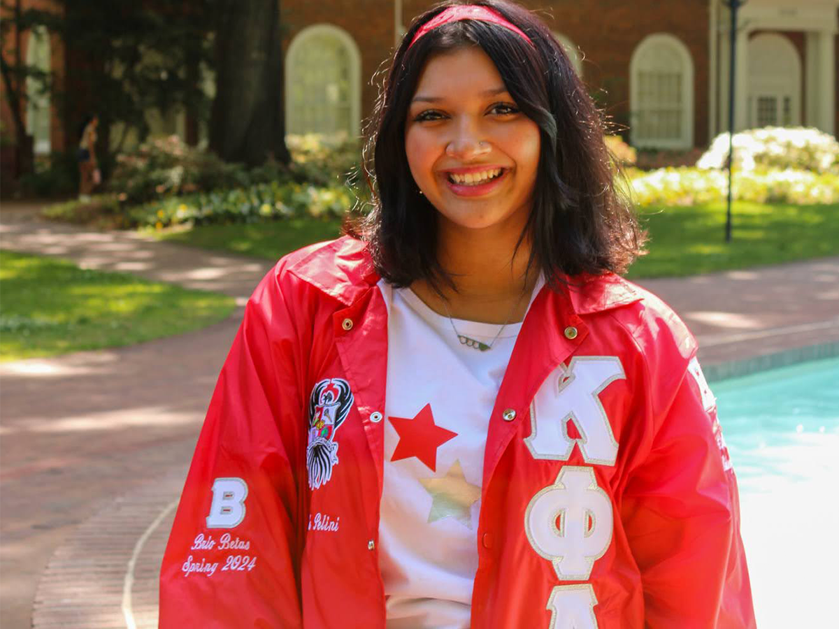 This photo features a young woman smiling warmly while standing outdoors near a fountain or water feature. She is wearing a bright red jacket with Greek letters ΚΦΛ (Kappa Phi Lambda) on the right side and various patches on the left, indicating sorority affiliation. One patch reads 