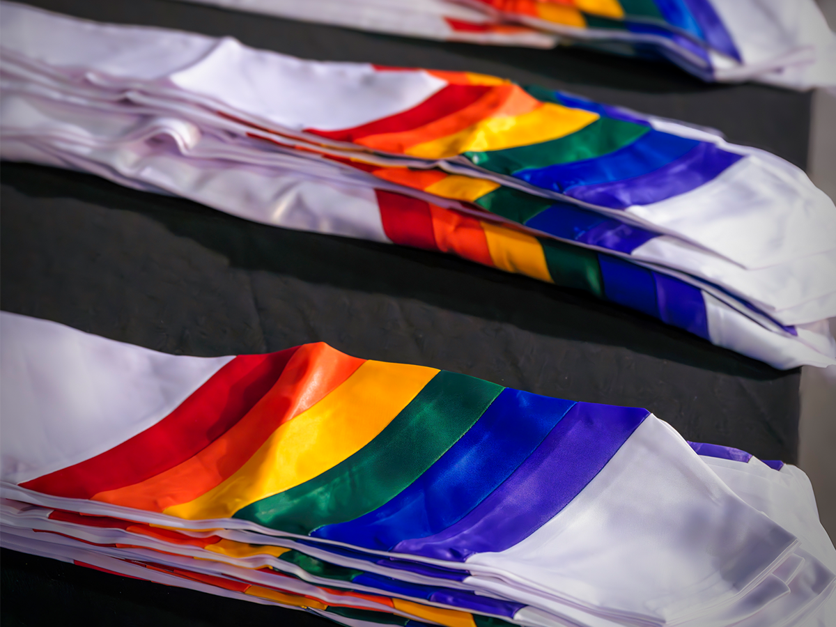 The photo shows a neatly arranged stack of white stoles with rainbow-colored stripes, laid out on a dark surface, likely a table. These stoles appear to be graduation stoles, often worn draped over the shoulders during commencement ceremonies. The rainbow pattern prominently represents LGBTQ+ pride, indicating that these stoles are likely for a Lavender Graduation ceremony—a celebration that honors LGBTQ+ students and their achievements. The vivid colors against the clean white fabric create a visually striking and meaningful display.