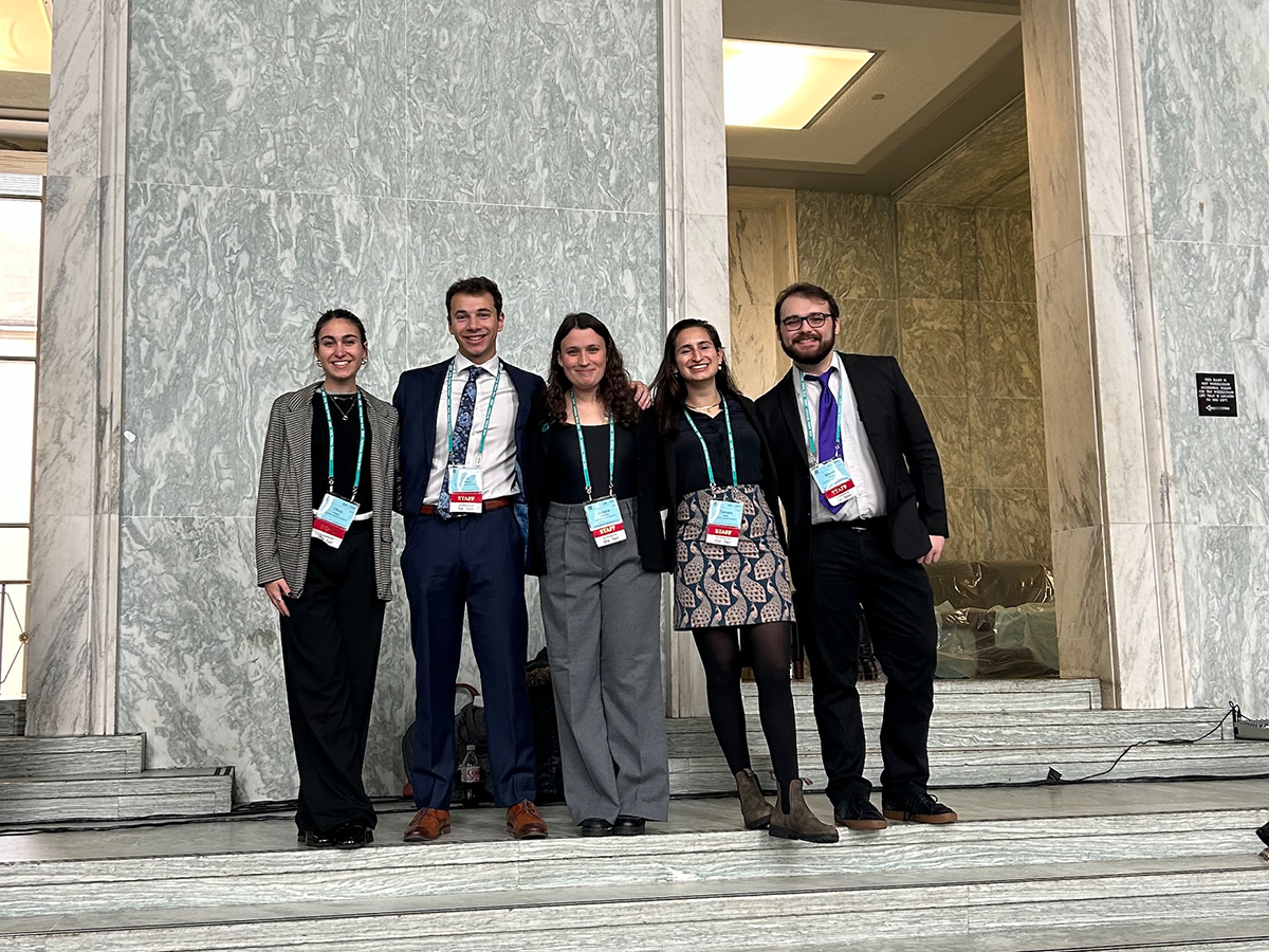 This photo shows five individuals standing together on a set of marble steps in what appears to be a grand, formal building—likely a government or institutional space, given the classical architecture, marble finishes, and prominent door frames. The group appears to be dressed in business or business-casual attire, possibly attending a conference or advocacy event, as suggested by the visible lanyards and name badges they are all wearing. The atmosphere is professional and collegial, and the people are smiling, suggesting a positive or celebratory moment.