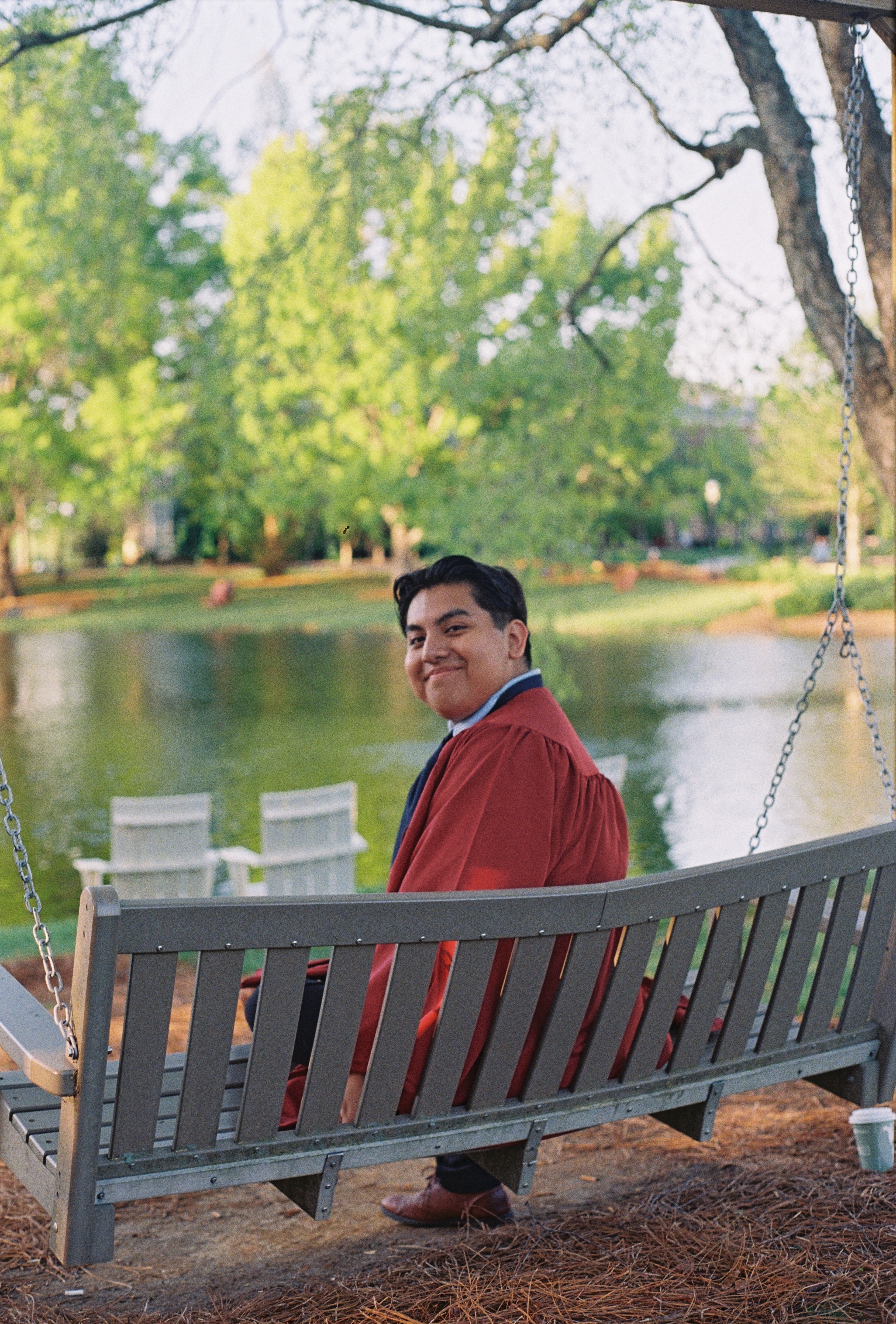 A graduate in maroon regalia smiles while sitting on a wooden swing by a peaceful lake, with spring trees and Adirondack chairs in the background.