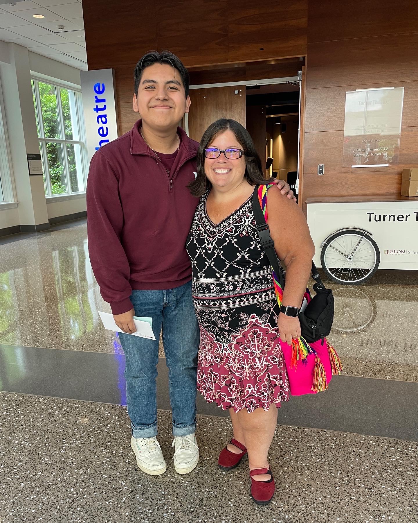 A smiling young man stands beside a woman in a patterned dress and bright accessories inside a modern campus building, both posing happily for the photo.
