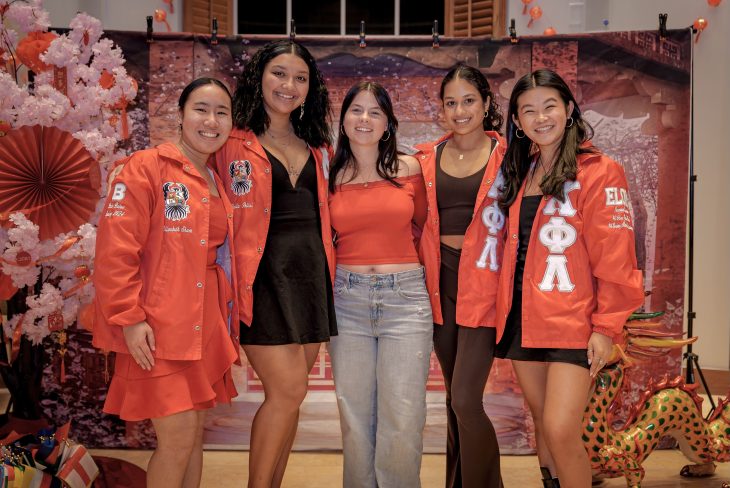 This photo shows a group of five young women posing together and smiling in front of a festive backdrop. The setting appears to be a celebration, likely for Lunar New Year, based on the decorations which include: Cherry blossom branches Red lanterns Red and gold paper fans A green and red dragon figure in the bottom right corner A backdrop that resembles traditional East Asian architecture and cherry blossoms Three of the women are wearing matching bright red jackets with Greek letters (suggesting a sorority or cultural organization), custom embroidery, and individual names or initials. One woman in the center is wearing a red off-shoulder top and light blue jeans, while another woman is wearing a black dress and a red jacket. The atmosphere is joyful and celebratory, with cultural pride and community spirit likely being key themes of the event.