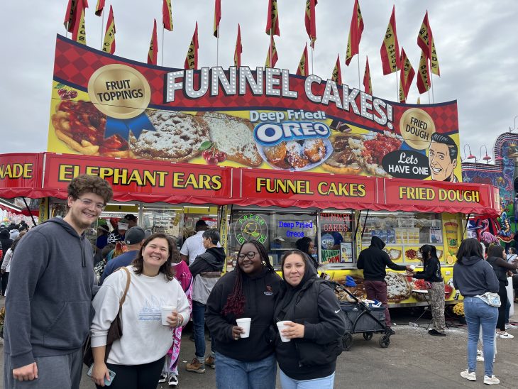 Four friends smile in front of a colorful fair stand selling funnel cakes, elephant ears, and deep-fried Oreos at a crowded outdoor event.