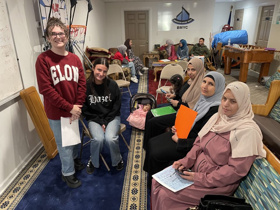 Two students, one wearing an Elon sweatshirt, smile beside a group of women in hijabs seated with papers and a baby in a community center, suggesting a cross-cultural educational or outreach activity.