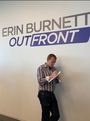 A man stands writing in a notepad beneath a large wall sign that reads “Erin Burnett OutFront.”