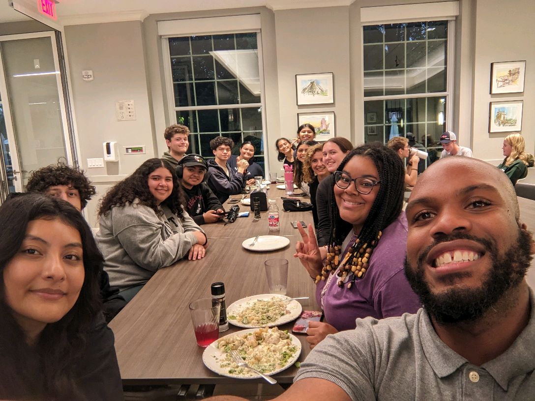 A group of students and a faculty or staff member smile for a selfie while gathered around a long dining table with plates of food in a brightly lit room.