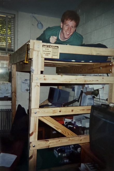 A smiling young man lies on a top bunk bed in a dorm room, with a cluttered desk and old computer setup below him.