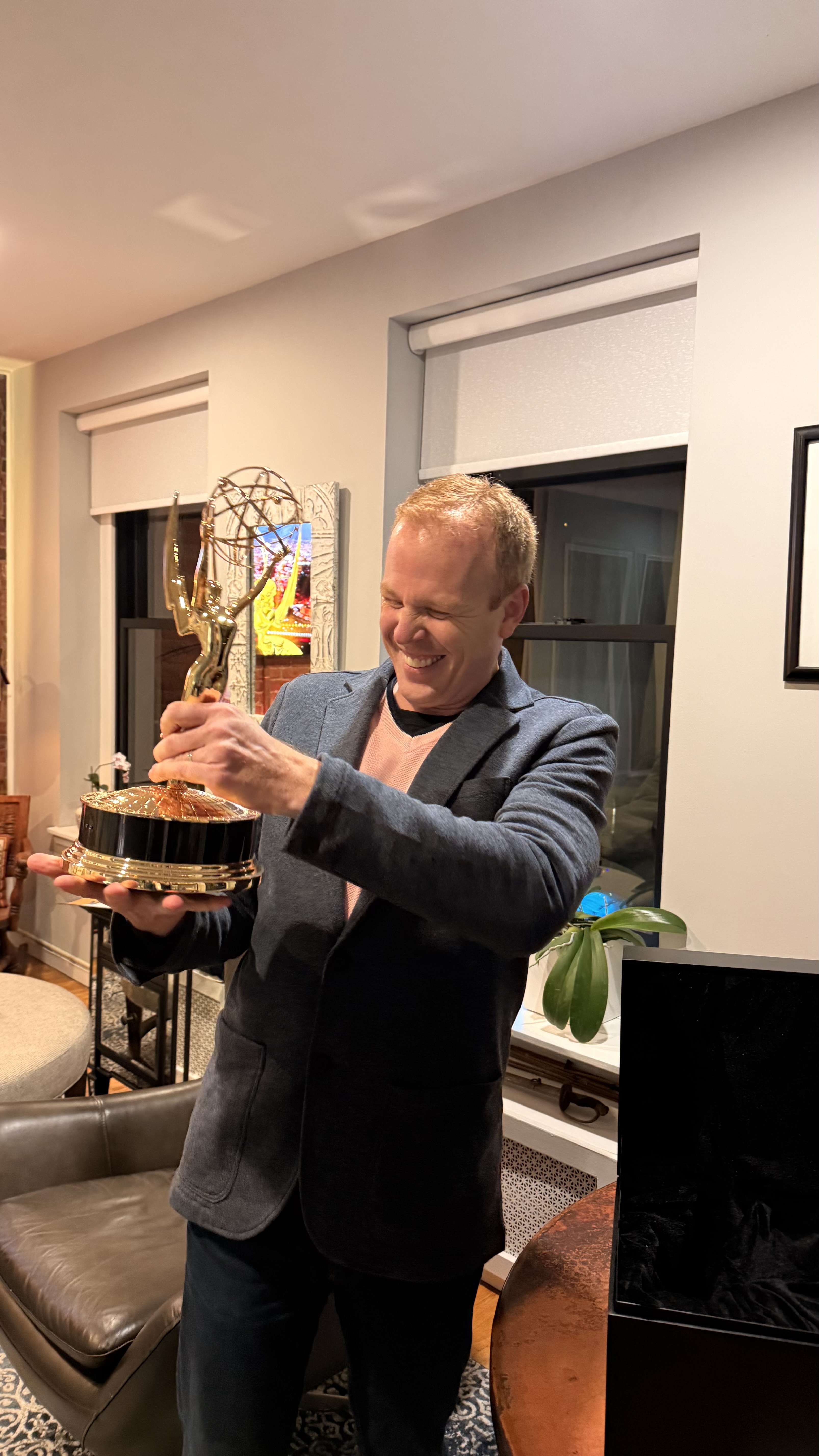 A man smiles joyfully while holding an Emmy Award trophy in a warmly lit living room.