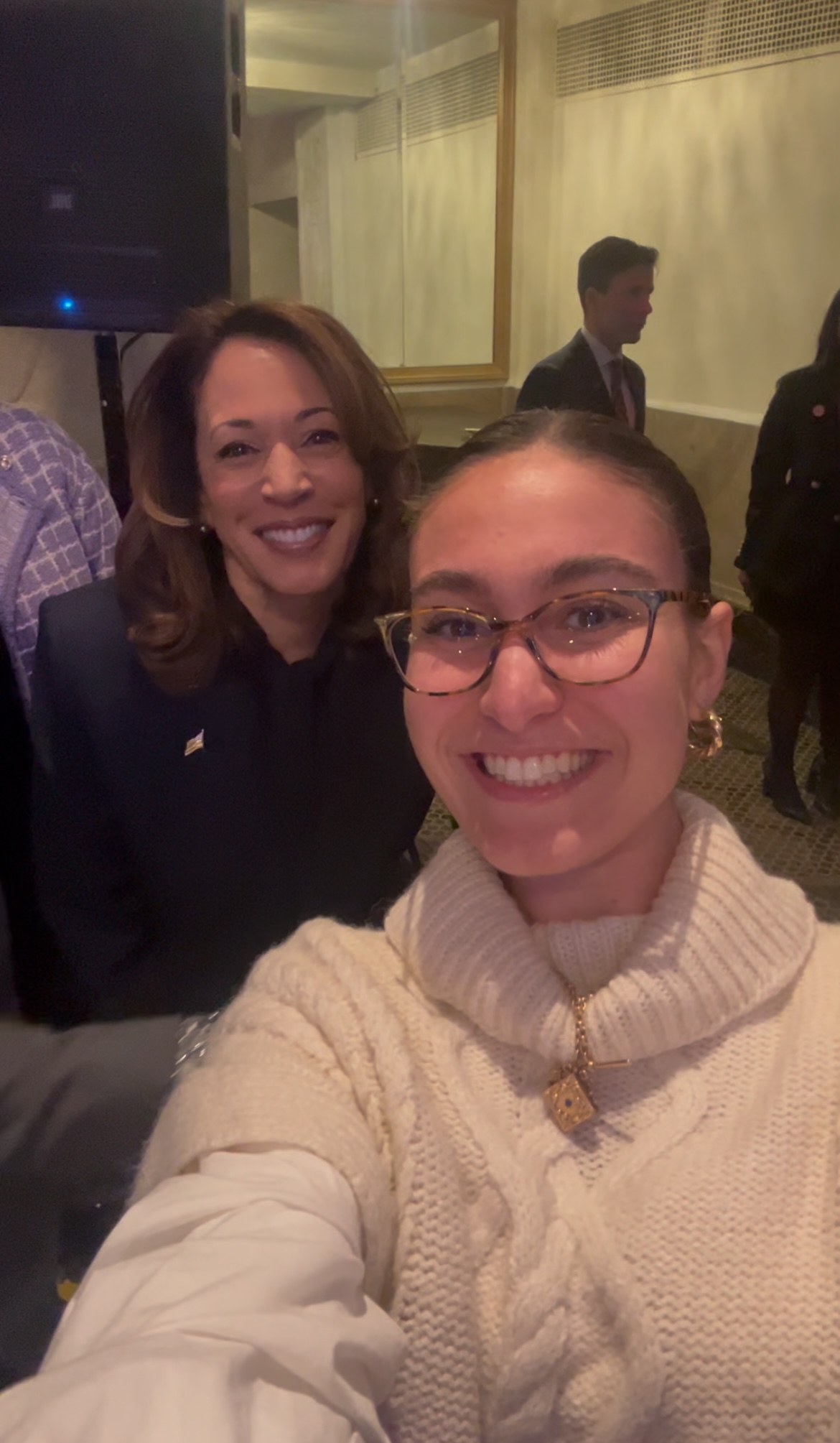 This photo shows two women smiling for a selfie in what appears to be an indoor event or formal gathering. The woman in the foreground, holding the camera, is wearing glasses, hoop earrings, and a thick cream-colored cable-knit turtleneck sweater with a distinctive gold necklace that features a pendant shaped like a padlock. The woman next to her, slightly in the background, is wearing a dark suit and has shoulder-length hair. There are other people in the background, and the lighting suggests a warm, possibly evening setting.