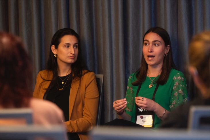 This photo shows two women seated and engaged in a discussion or panel talk in a professional setting. The woman on the right, wearing a green floral blouse and a name badge that reads "Olivia," appears to be speaking, gesturing with her hands as she explains something. The woman on the left, dressed in a tan blazer and black top, is listening attentively. Both women are wearing jewelry and look composed and professional. In the foreground, the backs of audience members' heads are visible, indicating this is likely a conference or seminar event. The background features dark blue curtains, adding to the formal ambiance.