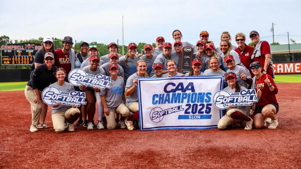 The Elon softball team poses together on the field holding signs and a banner celebrating their 2025 CAA Championship victory.