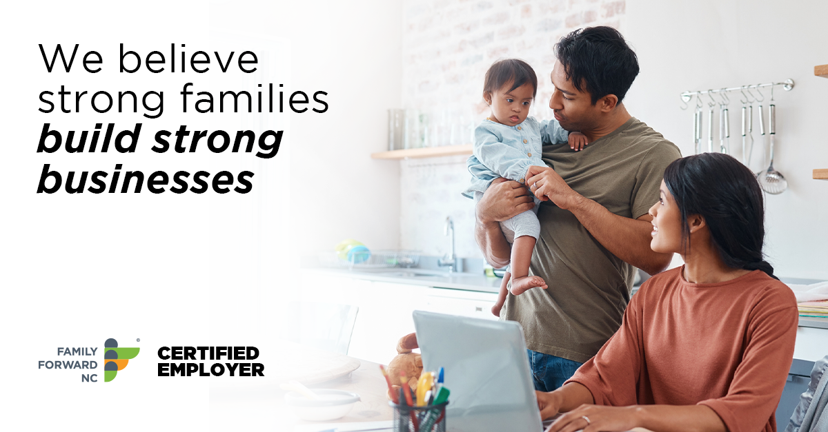 A banner of a father holding an infant while standing. The mother looks on from the table with her computer in front of her. Text over the photo reads "We believe strong families build strong businesses."
