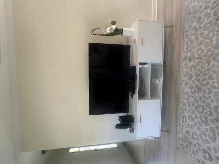 A flat-screen TV sits on a modern white media console with decorative items and electronics, next to a light-colored patterned rug on wood flooring.