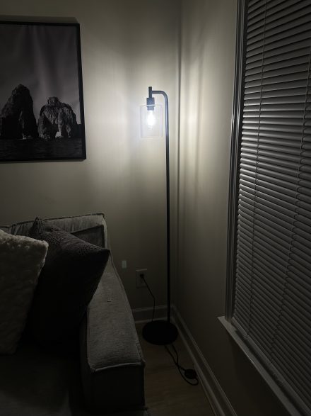 A lit floor lamp with a clear glass shade softly illuminates the corner of a room beside a gray sectional sofa and a window with closed blinds.