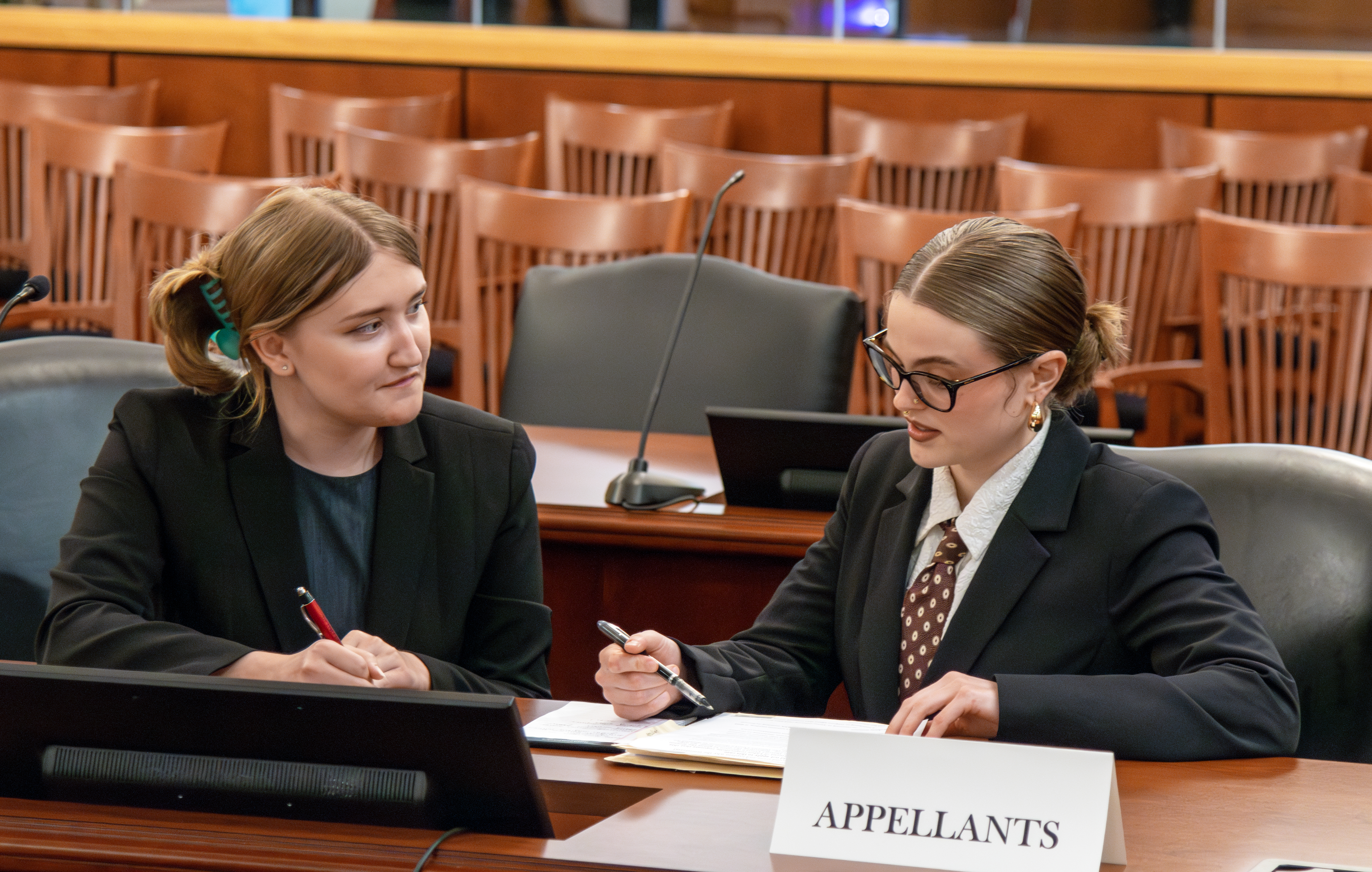 Two female law students in business attire seated at counsel's table in a courtroom. They are reviewing notes and conversing.