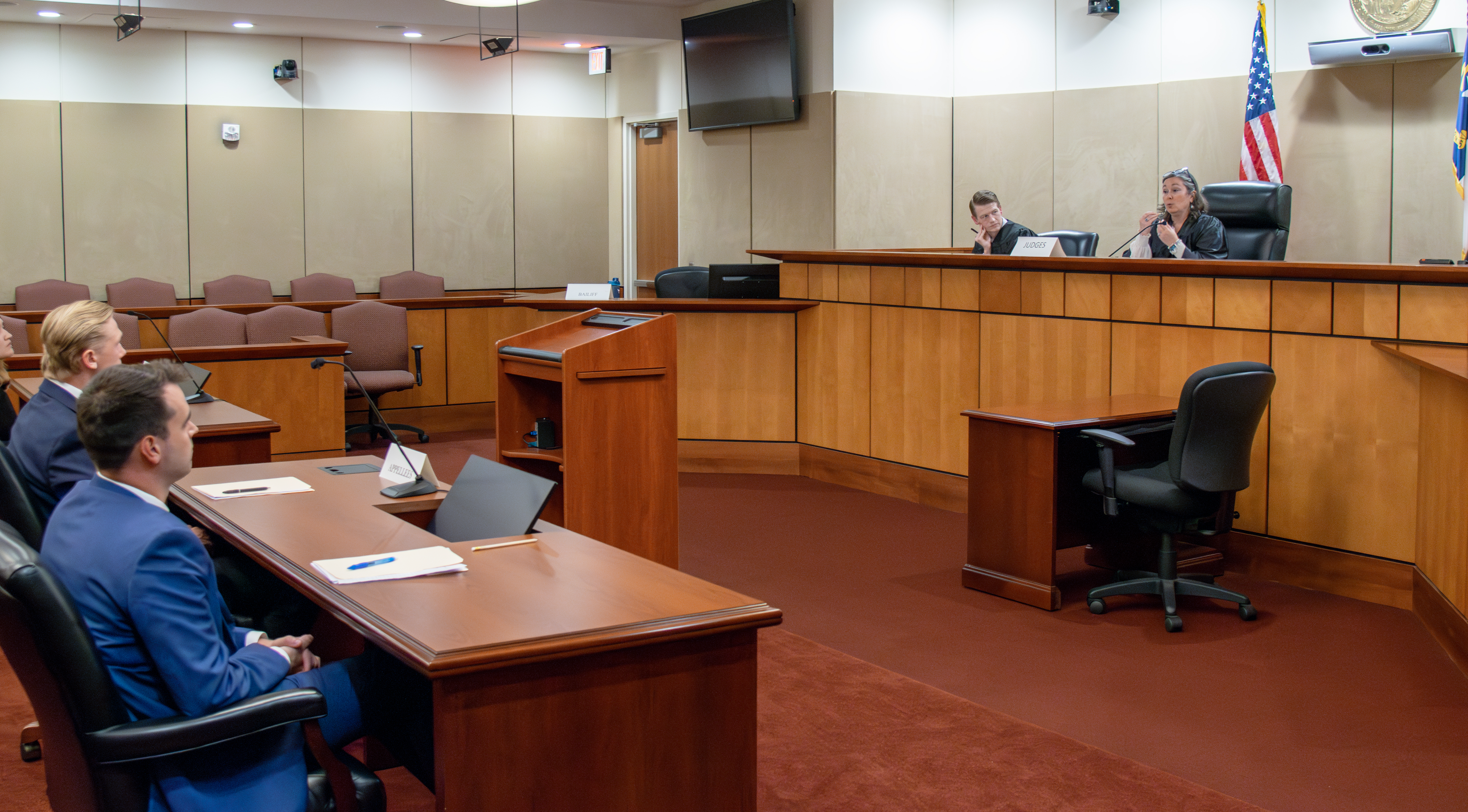 Two judges seated at the bench in a courtroom. A female judge is speaking. Two male students are seen listening from the counsel's desk.