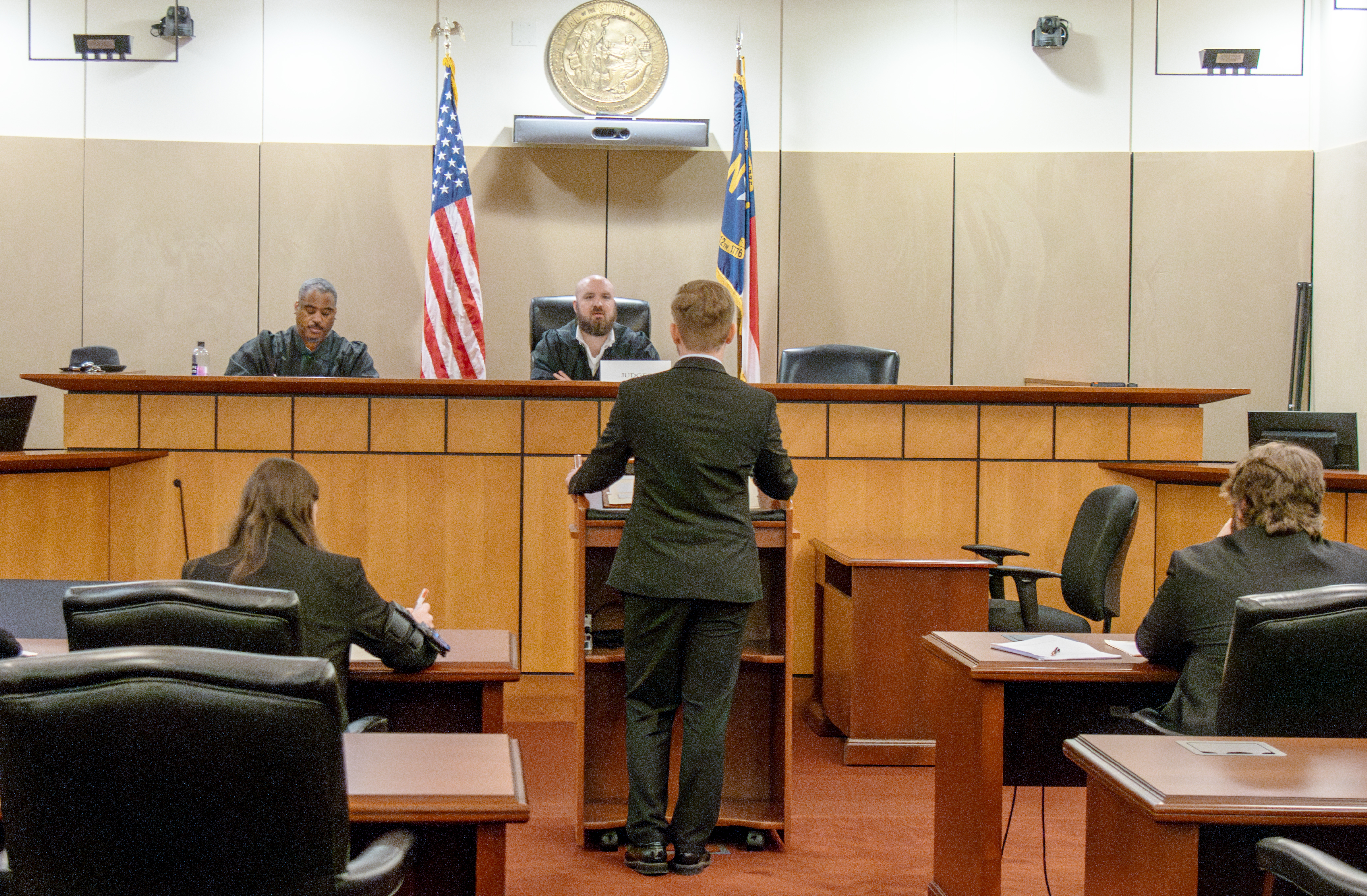 A view from the back of the courtroom. A male law student speaks at a podium before two judges who are listening and taking notes. Other law students are seated at counsel's tables on the left and right.