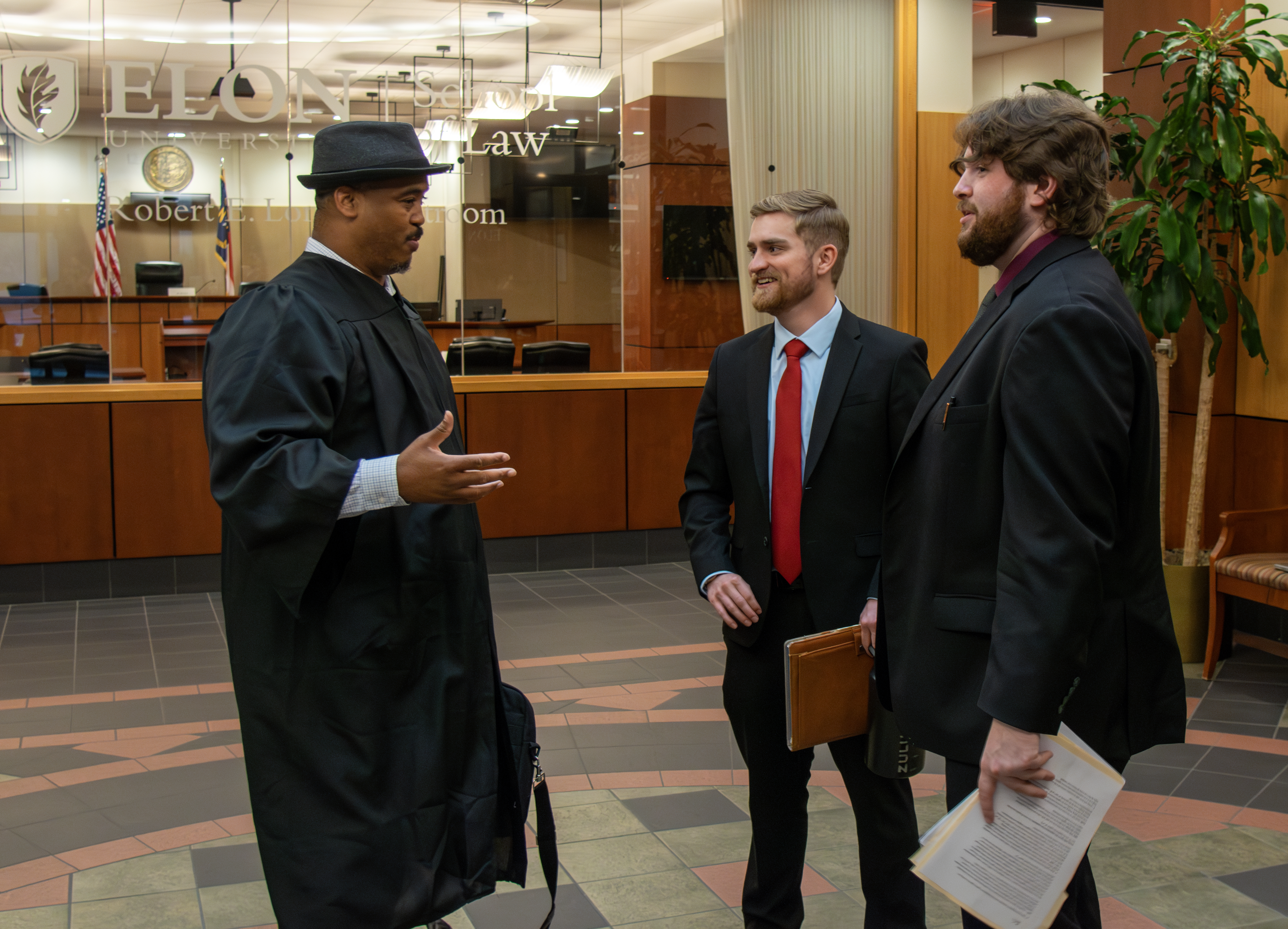 Three men in suits converse in the lobby of a law school. A courtroom is seen through a window behind them. They are smiling and engaged.