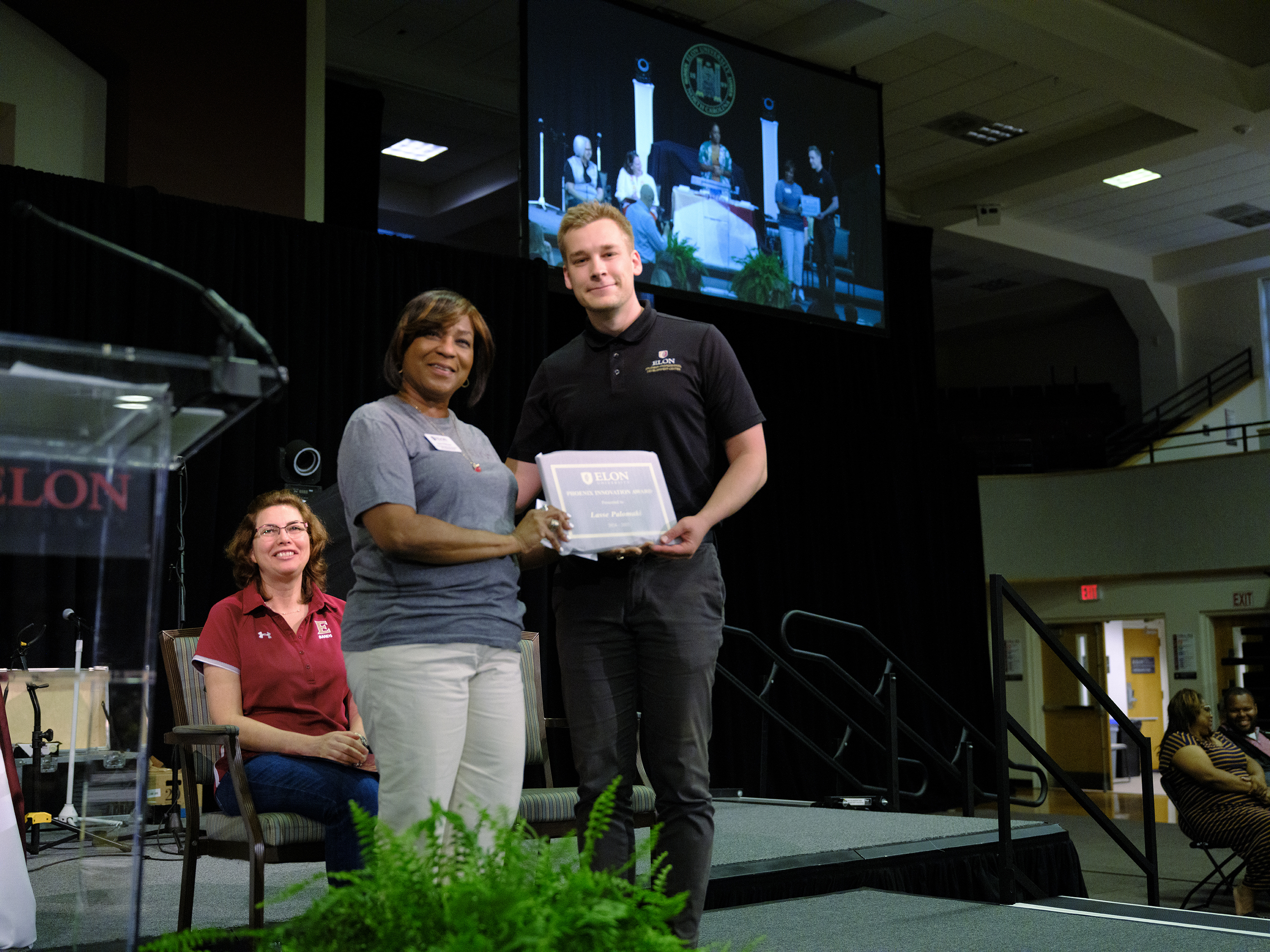 A woman and a man stand on stage holding a certificate together during an award ceremony, while another woman sits and smiles nearby.