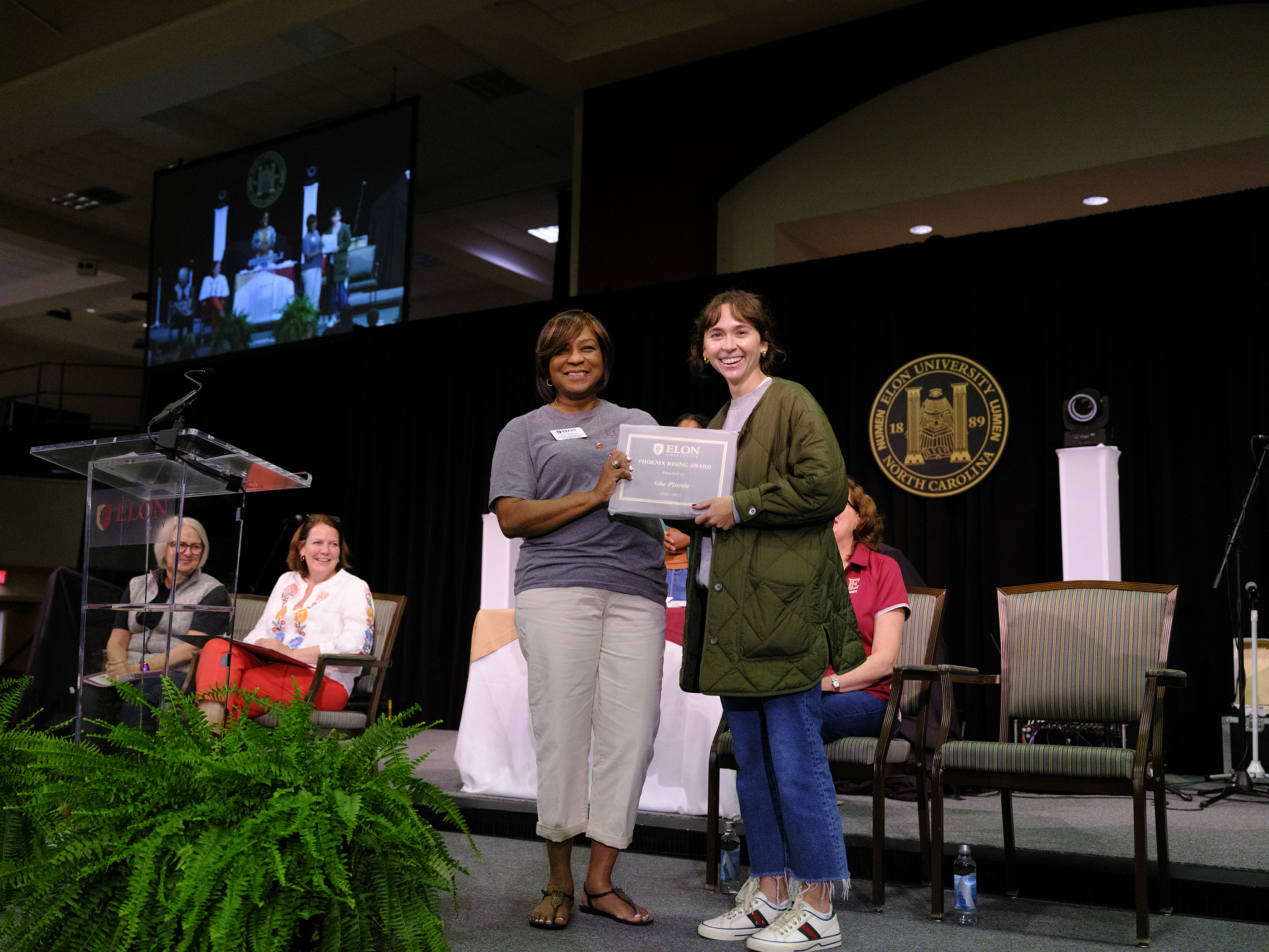 A smiling person receives a framed certificate from a woman on stage during an Elon University awards ceremony, with applauding attendees seated nearby.
