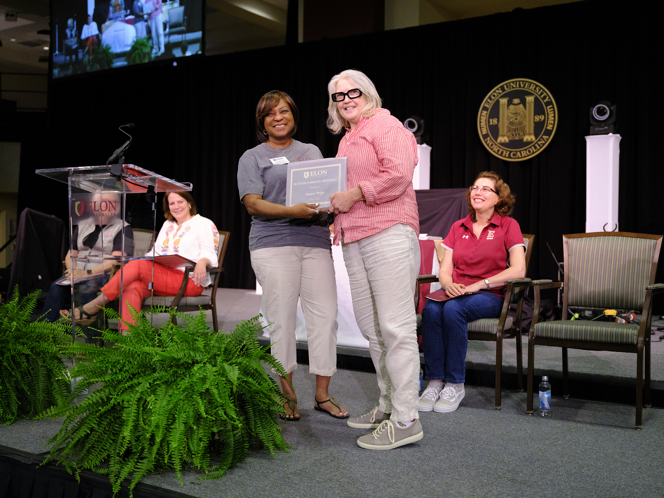 A smiling woman receives a framed certificate from a presenter on stage at an Elon University ceremony, as seated attendees watch and applaud.