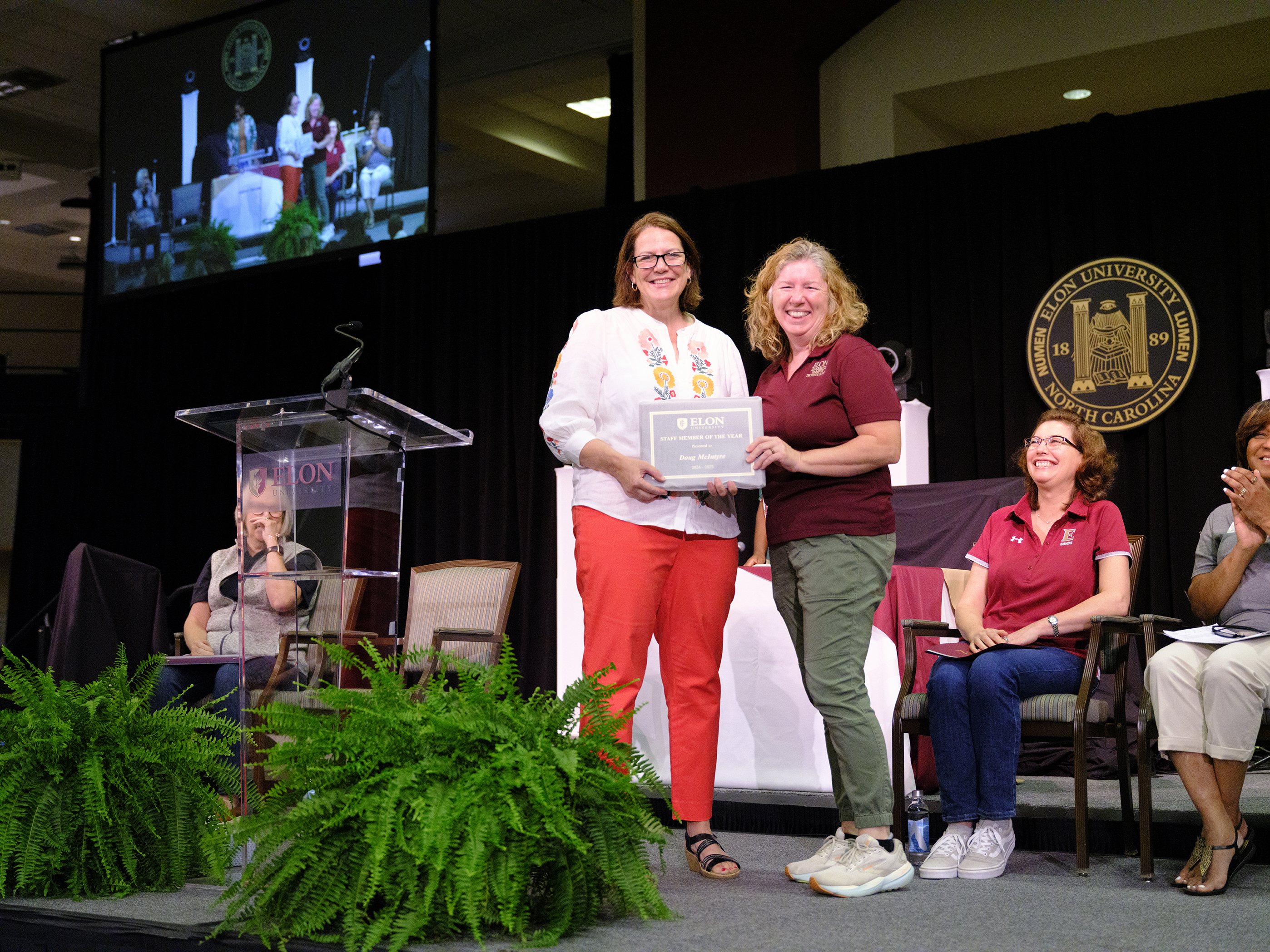 Two women stand smiling on stage holding a certificate at an Elon University ceremony, with colleagues seated and applauding in the background.