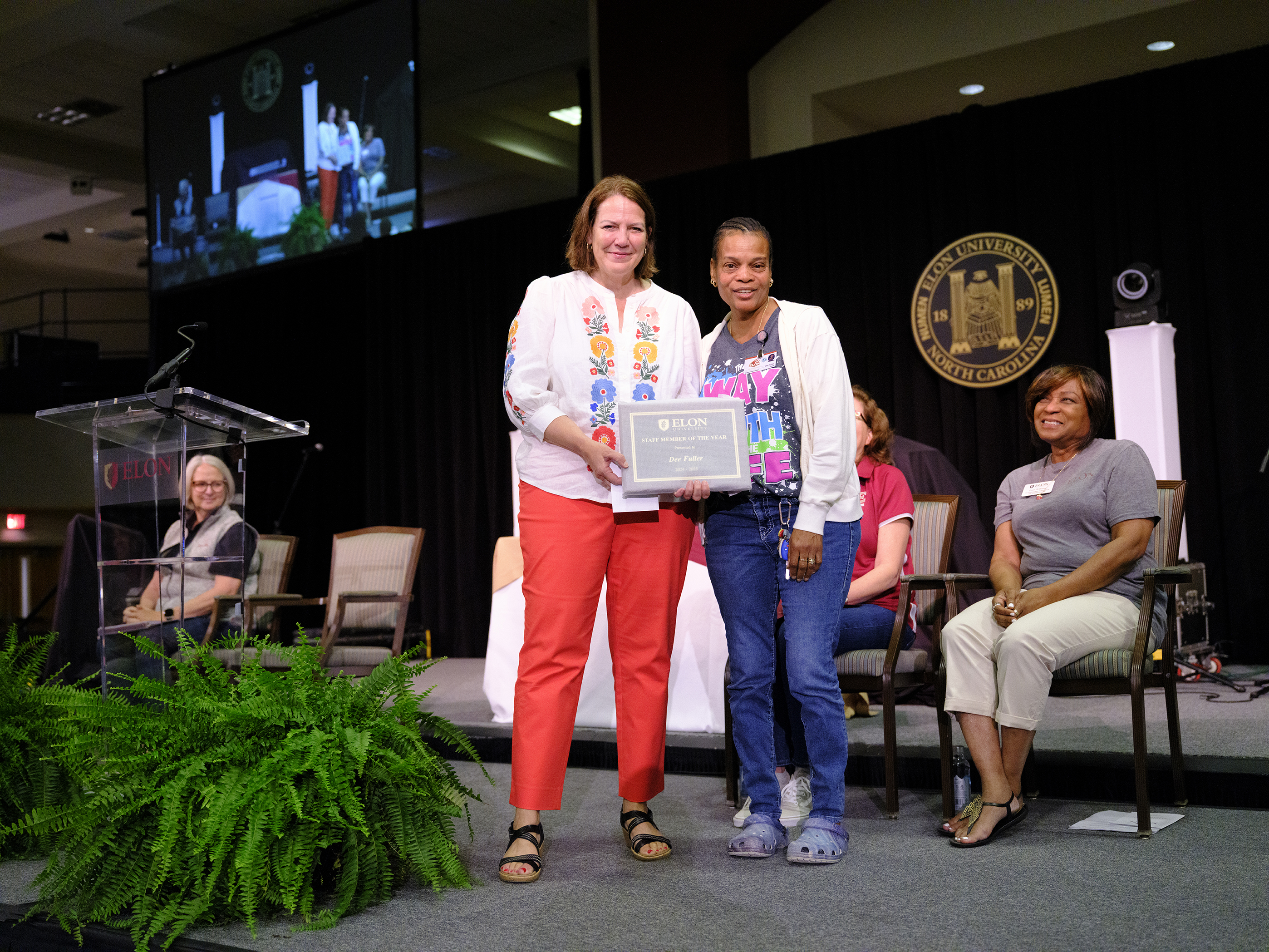 Two women stand at center stage smiling and holding a certificate during an Elon University award presentation, as seated colleagues look on supportively.