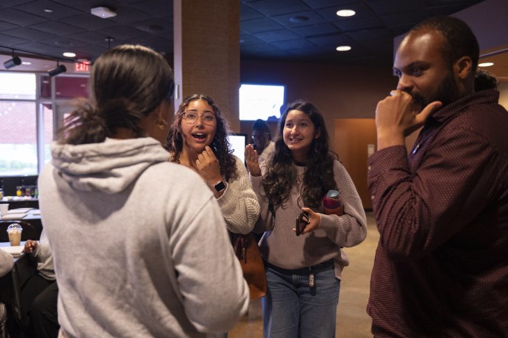 A group of four people engage in an animated and cheerful conversation indoors, with two women at the center smiling and gesturing enthusiastically.