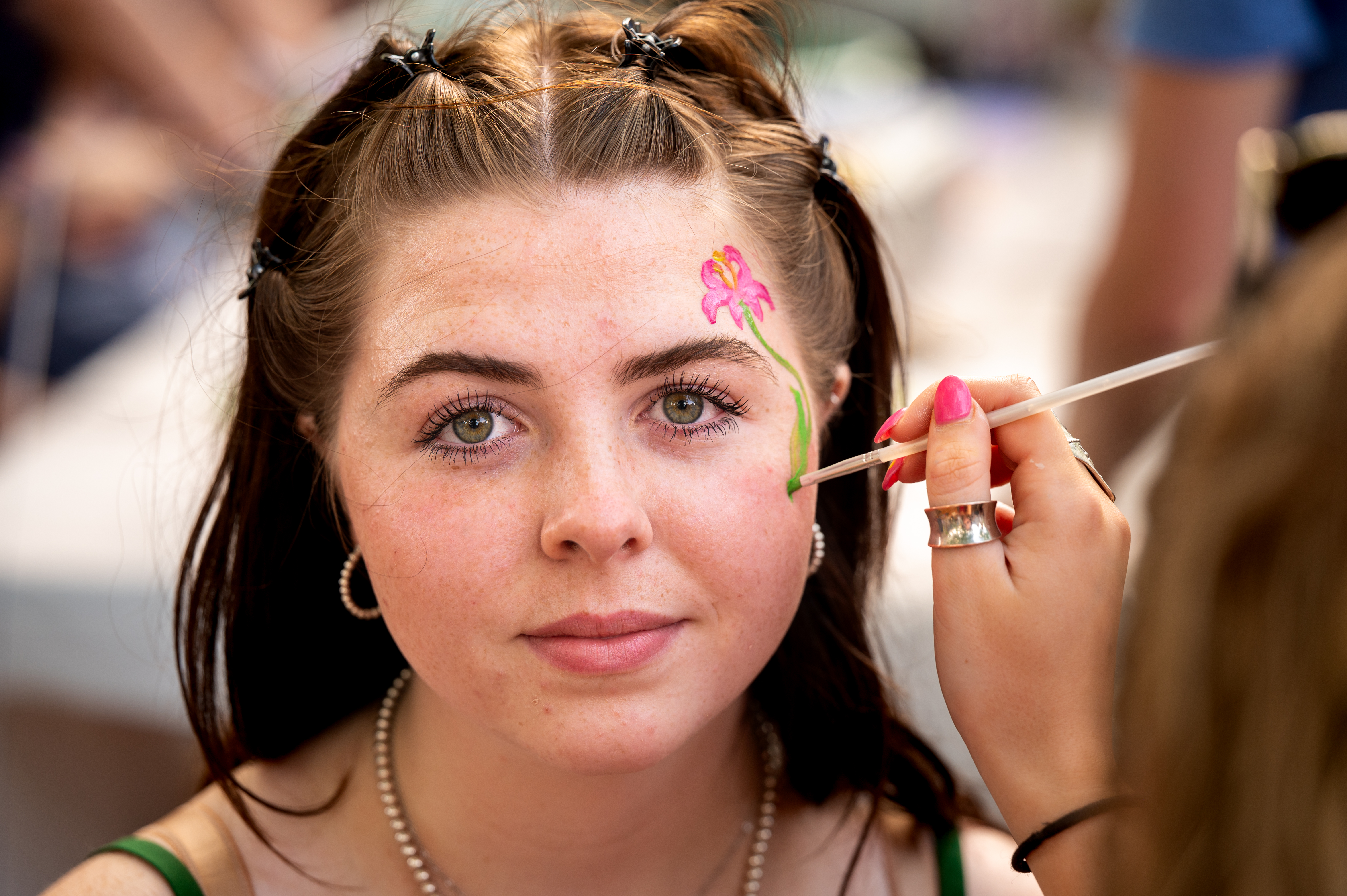 A young woman with pearl earrings and a necklace smiles slightly as another person paints a pink flower on her face at a face-painting event.