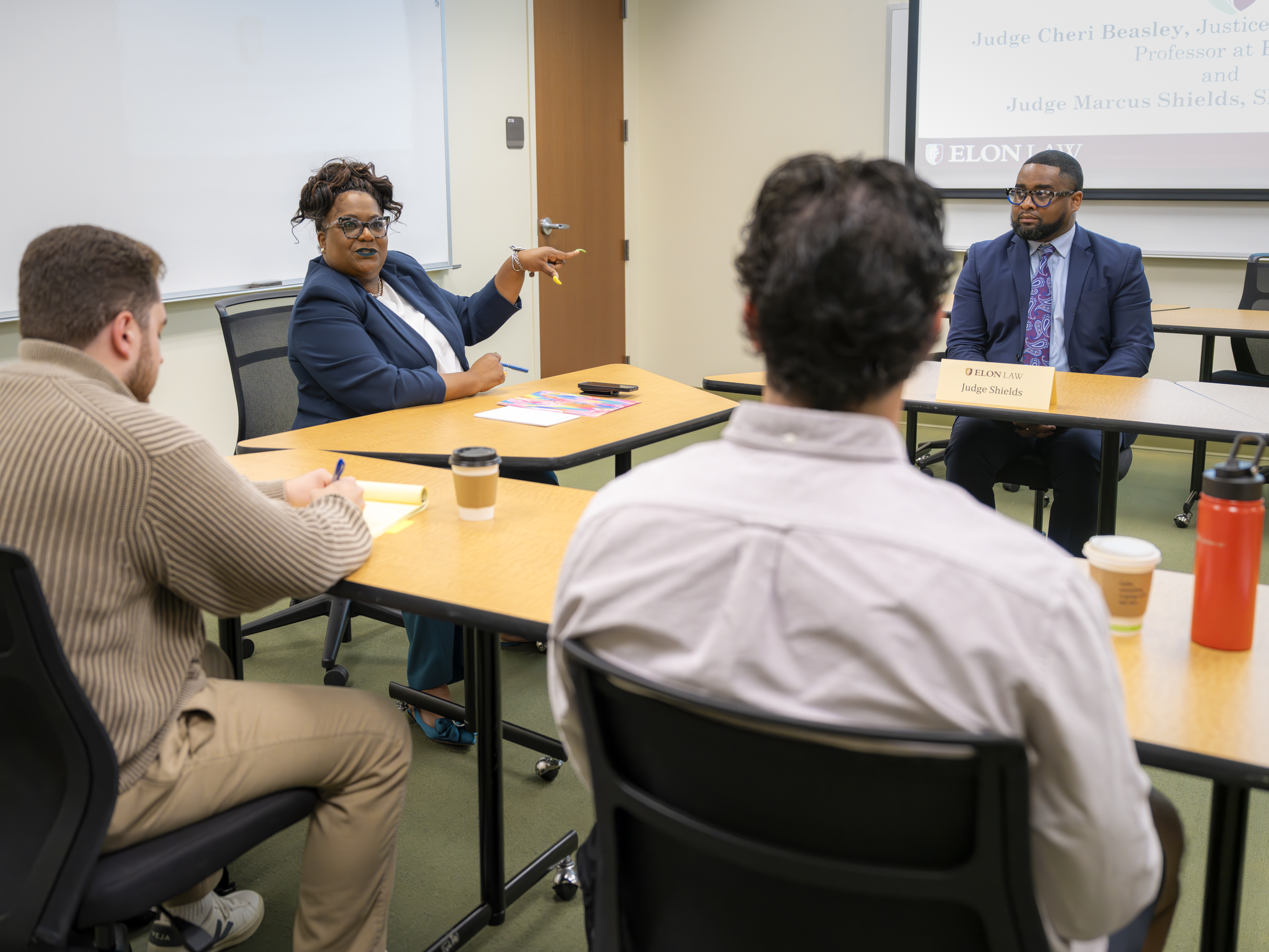 A wide view of a class in a roundtable discussion with a professor gesturing toward a guest speaker.
