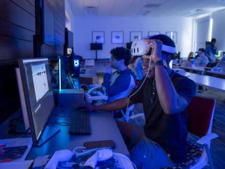 A student lifts a VR headset from his eyes while working at a computer in a dimly lit classroom, surrounded by classmates engaged in virtual reality programming.