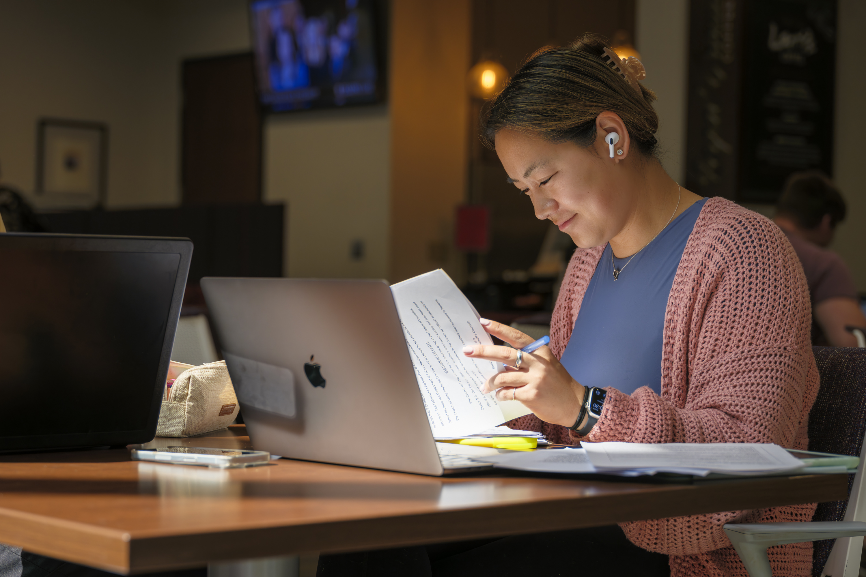 A student wearing wireless earbuds studies at a table with a laptop and printed documents, highlighting text while smiling in a warmly lit indoor space.