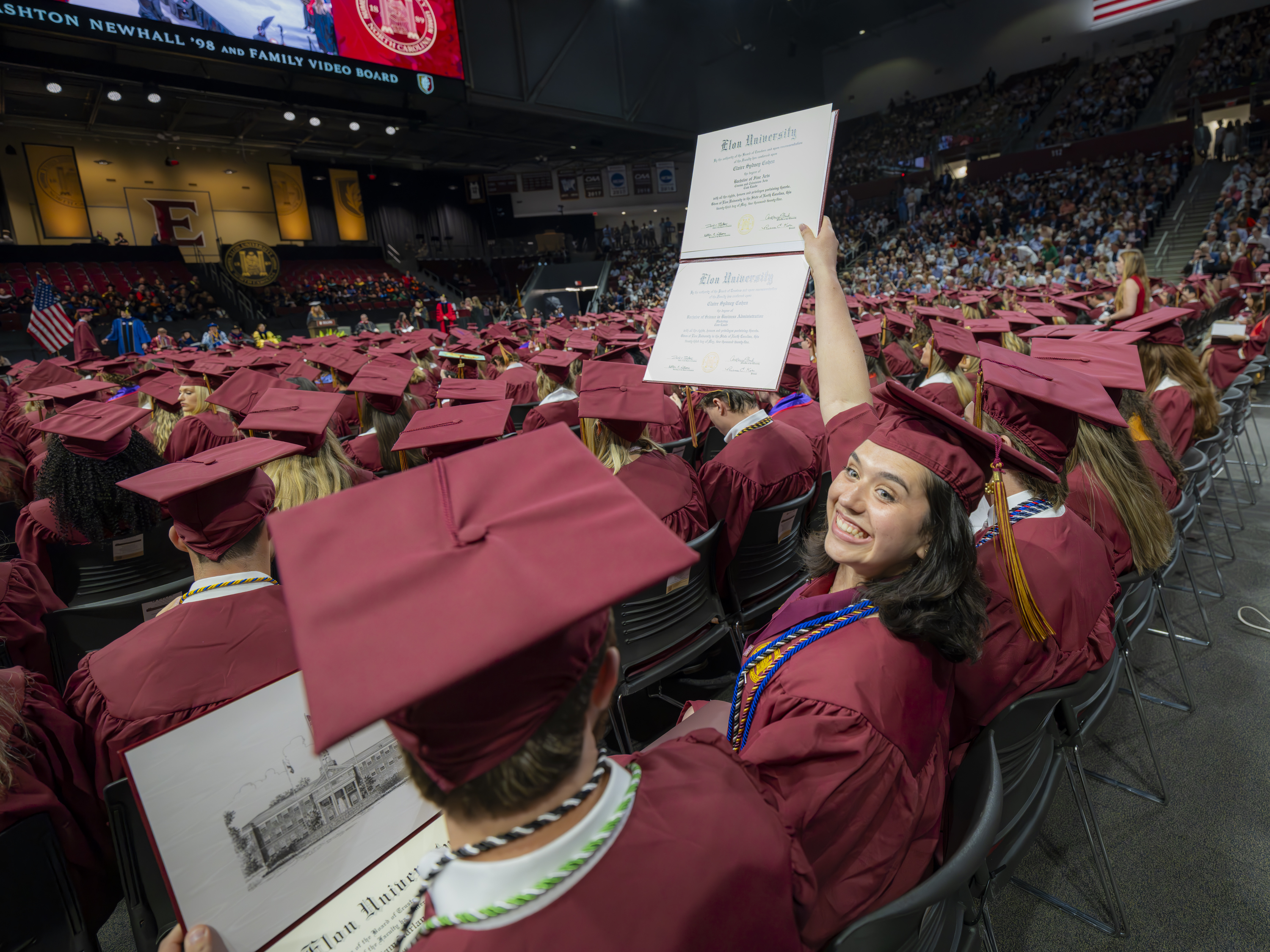 A joyful graduate in a maroon cap and gown smiles and raises her diploma high above her head while seated among a sea of fellow graduates at Elon University’s commencement ceremony.