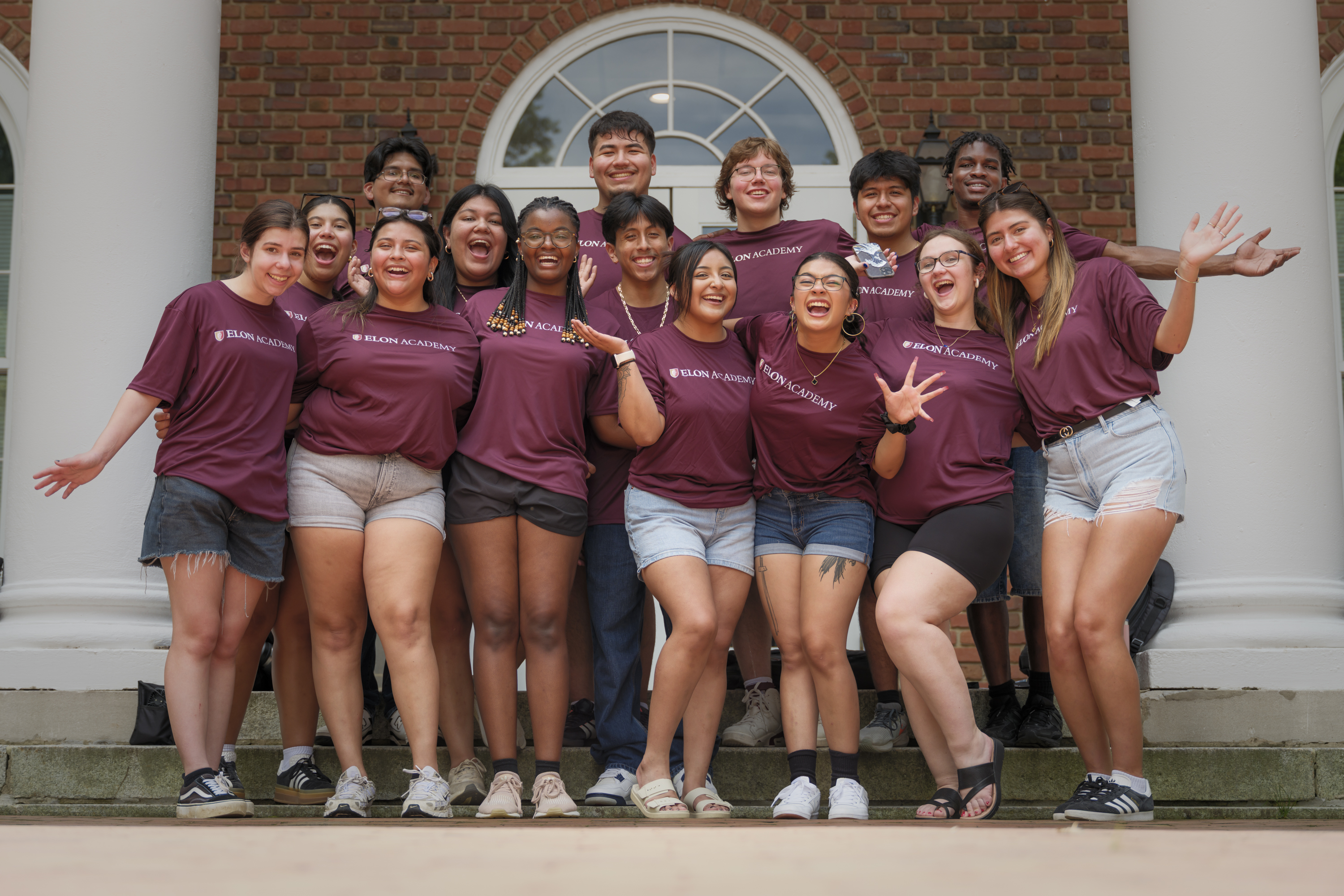 A group of smiling Elon Academy students in matching maroon shirts pose energetically together on outdoor steps in front of a brick building with white columns.