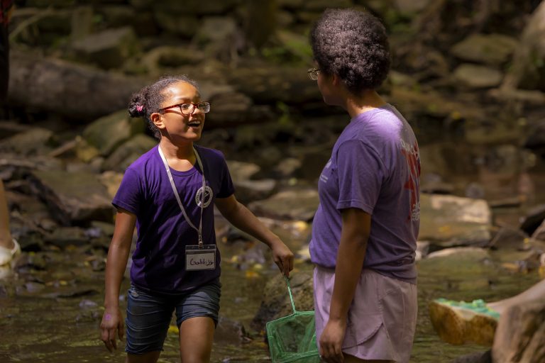A girl smiles while carrying a green net and talking with a friend in a creek.