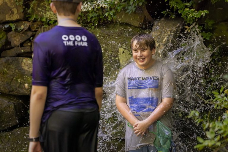 A smiling child stands under a small waterfall, enjoying getting splashed, while another person stands facing them.